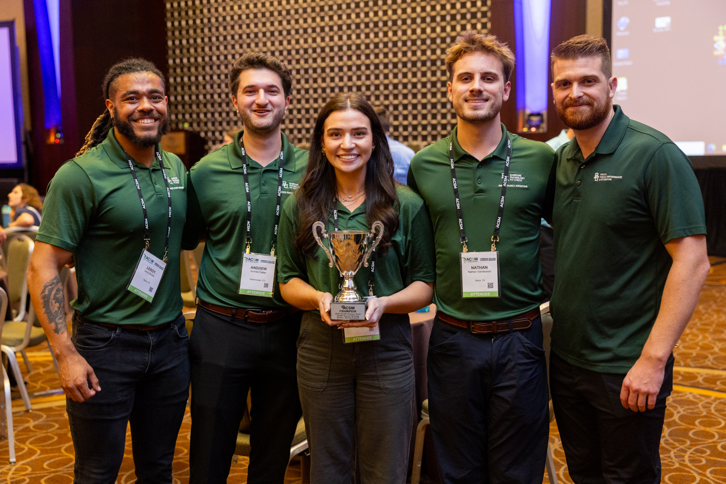 ACSM 2024 student bowl winners, team from Texas. four men and one woman all wearing jeans and dark green polos. the woman in the middle is holding a trophy